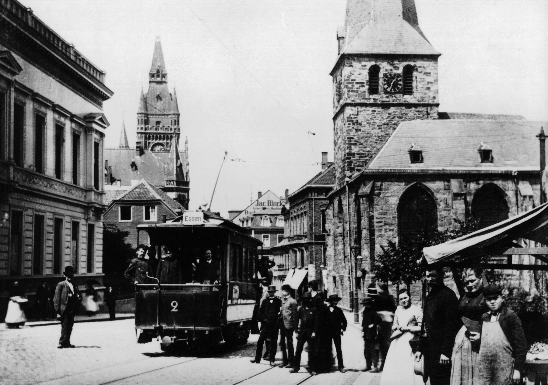 Die ersten Probefahrten der neuen Straßenbahn auf der Kettwiger Straße waren 1893 erkennbar ein Ereignis für die Essener. Unverändert ist die Johanniskirche geblieben, die das Wiedererkennen erleichtert, alle anderen Gebäude gibt es nicht mehr. Links ist der Turm des alten Rathauses zu sehen. Die Bahn hat einen hochmodernen elektrischen Stromabnehmer, die Leitung musste längs der Straße gespannt werden.