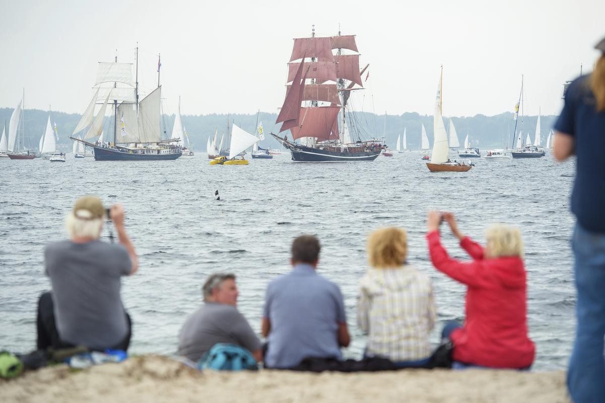 Menschen beobachten vorbeifahrende Segelschiffe während der traditionellen Windjammerparade zum Abschluss der Kieler Woche in der Kieler Förde vom Strand in Friedrichsort aus. 