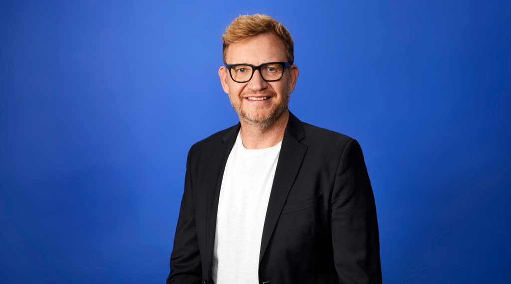 Karl Valks, der von Sky zur Sportschau geht, schaut lächelnd in die Kamera.