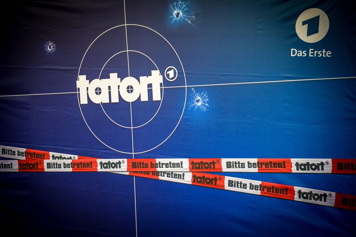 Auf dem Bild sieht man eine blaue Leinwand mit dem Tatortlogo in weißer Schrift drauf. Um das Wort "Tatort" sind zwei Kreise, welche aussehen wie eine Zielscheibe. Unten im Bild befinden sich ein weiß/rotes Absperrband mit der Aufschrift "Bitte Betreten, Tatort".