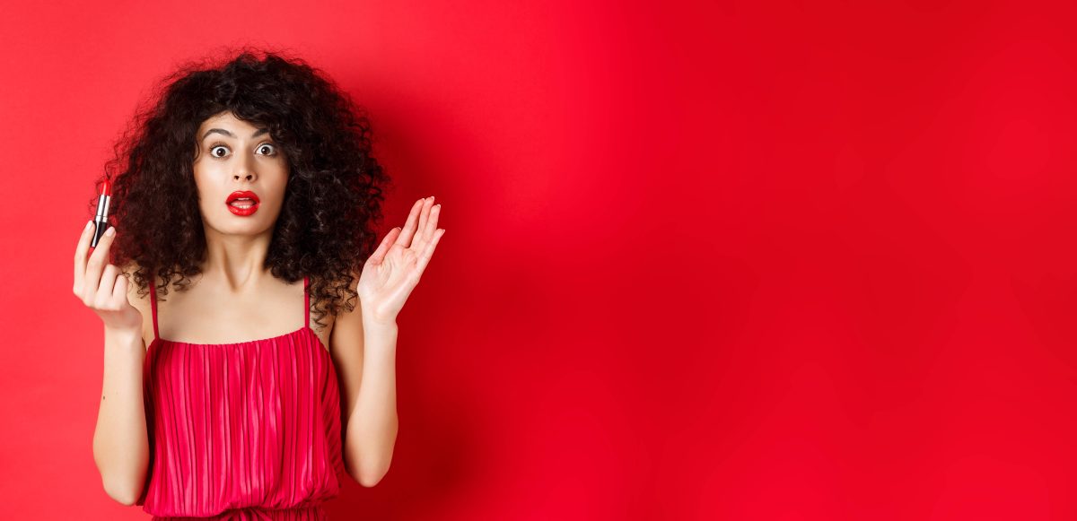 Das Bild hat einen roten Hintergrund, vor dem eine Frau steht. Die Frau trägt roten Lippenstift und ein rotes Kleid. Sie hat in ihrer linken Hand einen Lippenstift und schwarze lockige Haare. Sie hat ihren Mund auf und schaut erschrocken.