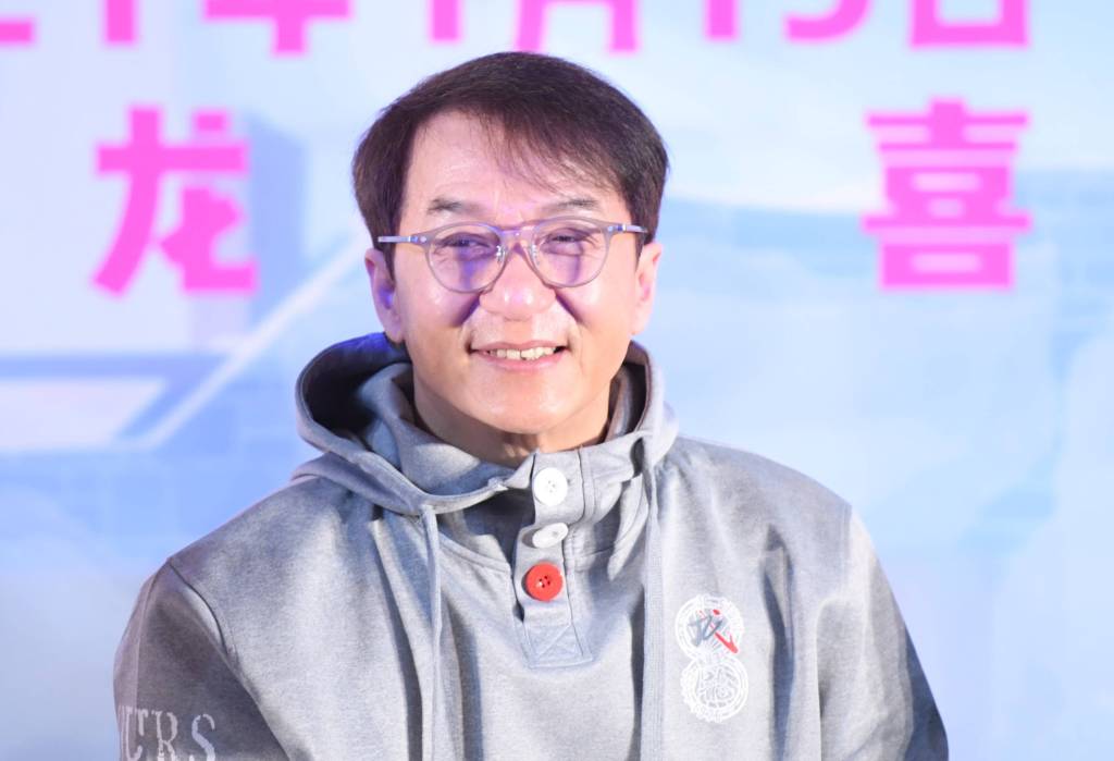 Jackie Chan, er trägt eine Brille und einen grauen Pullover.