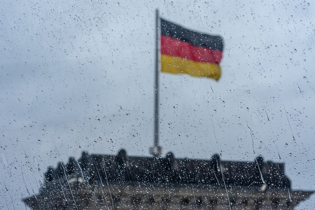 Eine verregnete Fensterscheibe vor dem Hintergrund einer Deutschland-Fahne, die im grauem Himmel weht.